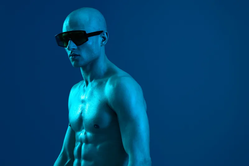 Athletic man with sunglasses posing against a vibrant blue background, showcasing strong physique and confidence. Perfect for fitness and lifestyle themes.