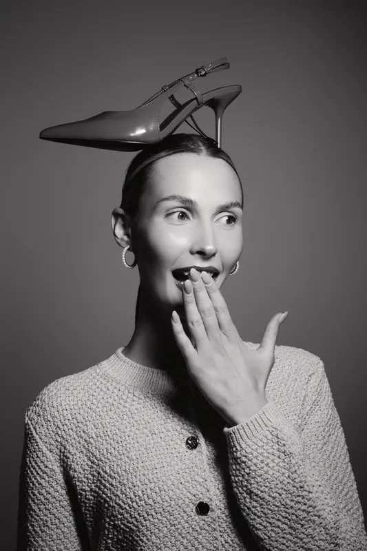 Young white woman with fair skin and earrings balances high heel shoe on head, hand over mouth in surprise, black and white portrait concept of fashion fun.