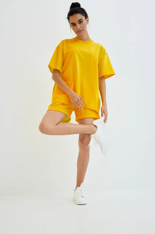 Young woman posing in bright yellow casual outfit with white sneakers, standing on one leg against plain white background, relaxed and modern style.