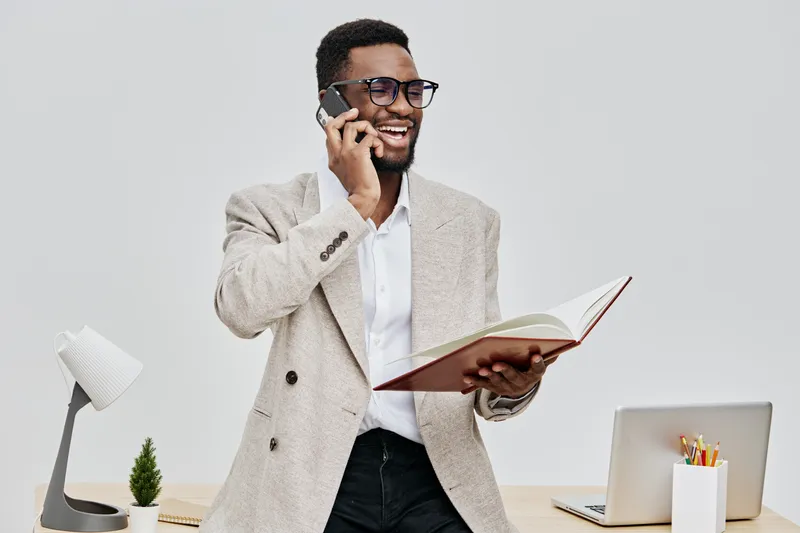 Smiling man with glasses talking on mobile phone while holding open notebook, dressed in beige blazer and white shirt, working in modern office with laptop and desk lamp, concept of business communica