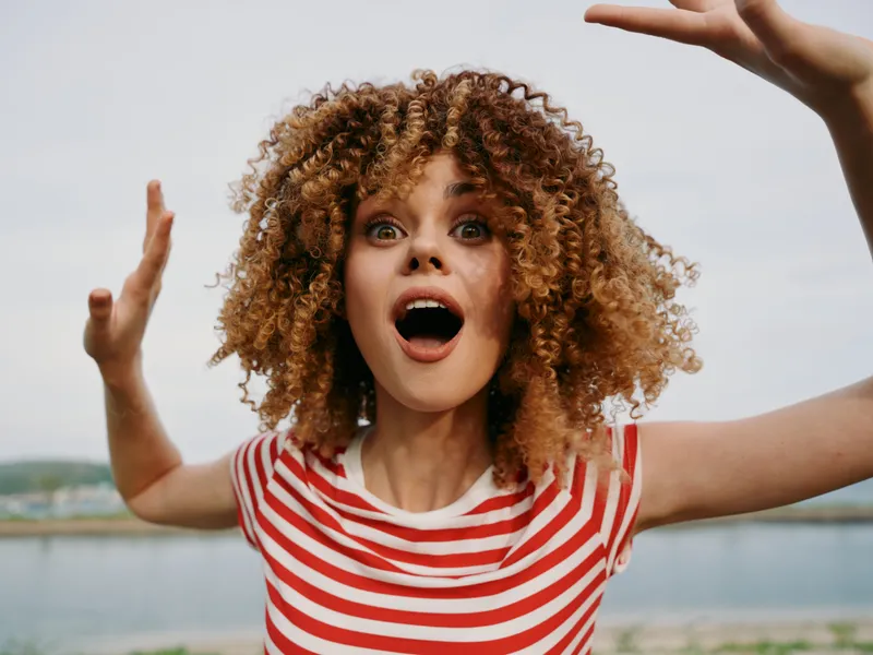 Young woman with curly hair in a red striped shirt expresses surprise with wide eyes and open mouth, arms raised, outdoors by a calm lake under bright sky.