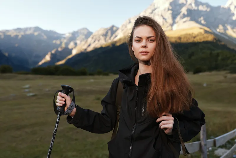 A resolute hiker stands in an alpine landscape, gripping a trekking pole as the wind brushes her hair, conveying resilience and adventure in natural surroundings.