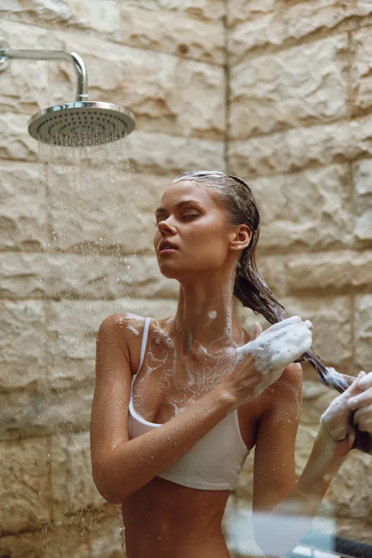 woman showering outdoors under rainfall, washing hair with soap foam, natural stone wall background, refreshing morning routine in warm sunlight