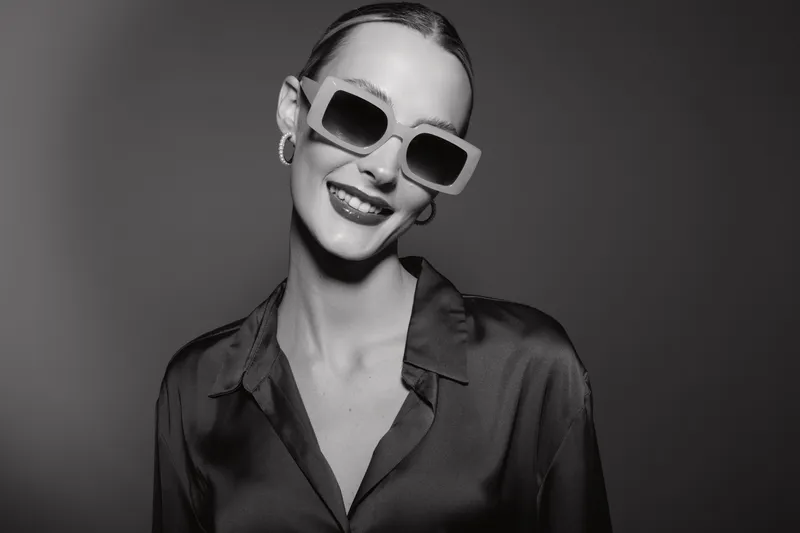 Smiling Caucasian woman with pale skin wearing large stylish sunglasses and hoop earrings poses confidently in a dark satin shirt on a plain background. Concept of fashion and elegance.