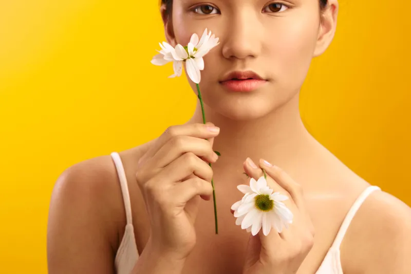 asian woman holding daisies against a yellow background, gazing softly at the camera. the simple composition conveys calm confidence, natural beauty, and a fresh, optimistic mood.