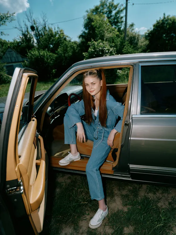 Young woman in casual outfit sitting in a car, smiling outdoors, with a bright blue sky and green trees in the background, showcasing a carefree lifestyle.