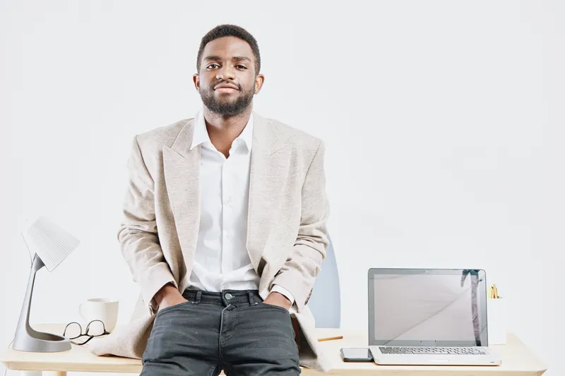 Confident young man with beard wearing beige blazer and white shirt standing in modern office with desk, laptop, paper, and glasses, looking at camera on plain white background. People lifestyle conce
