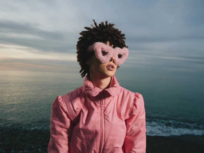 Pink masked portrait of a person standing by the sea in a pink shirt, sunset light, whimsical fashion accessory and ocean breeze.