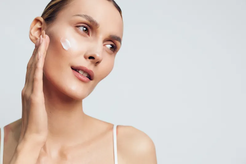 Young woman applying facial cream on clean skin, natural beauty concept, soft lighting, minimal makeup, smooth complexion, skincare routine, serene expression, light background