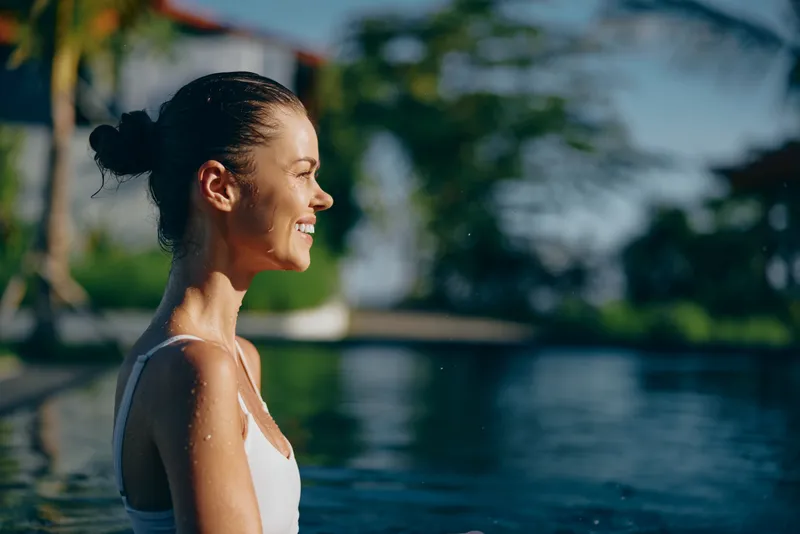 Smiling woman with hair tied in a bun standing by a sunlit pool, water behind. Casual summer lifestyle, wellness, outdoor relaxation, warm light, bright mood.