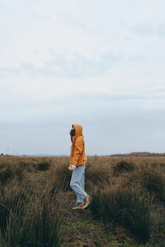 Woman lifestyle outdoors walking through grassy field wearing orange jacket and blue jeans, side view, soft daylight sky, calm mood, nature travel feeling, rural countryside, full body