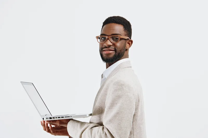 Professional smiling man with glasses, dark curly hair and beard, wearing light-colored blazer and white shirt, holding open laptop, looking at camera on plain white background, studio portrait, busin