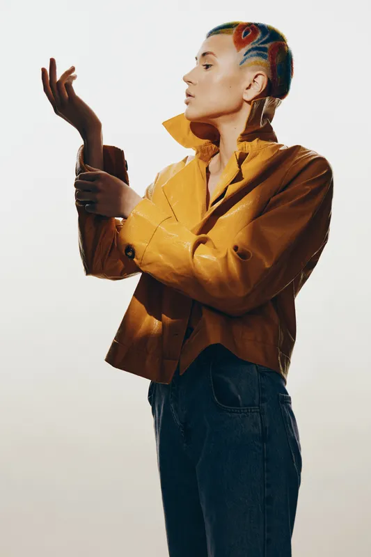 Woman with colorful shaved hairstyle wearing a mustard jacket and jeans, posing thoughtfully against a light background in stylish casual fashion.