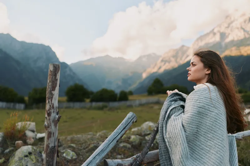 Young woman with long brown hair stands near a wooden fence in a mountain meadow, wrapped in a soft gray knit shawl, gazing into the distance with a peaceful, thoughtful mood.
