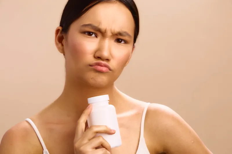 asian woman holding a white bottle, looking thoughtful and confident, indoors with a soft beige backdrop, health and wellness theme, product focus and care.