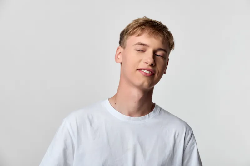 Young man in a plain white t shirt rests with eyes closed and a subtle smile, relaxed in a simple studio setting, conveying ease, calm, casual mood and gentle confidence