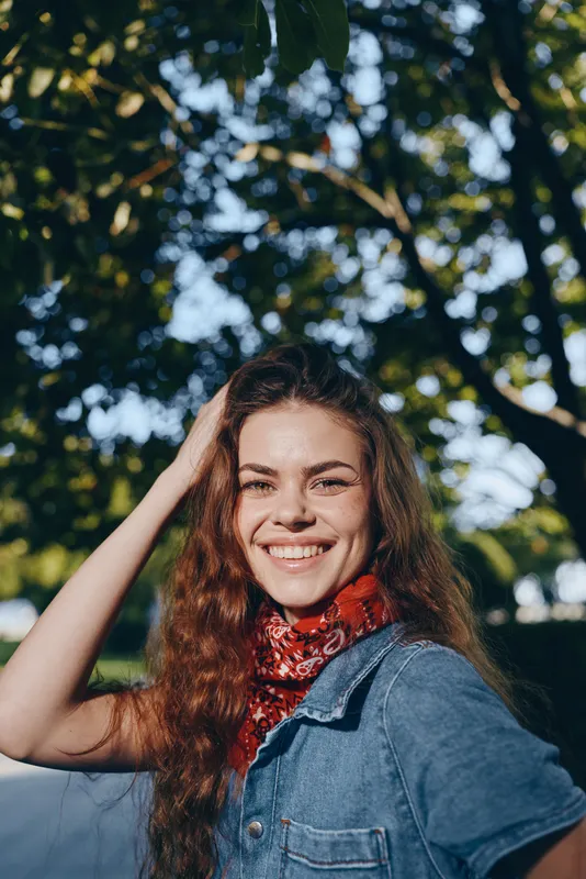 smiling woman in modern boho-western denim street style with film color effect, red bandana, natural light, and green foliage background outdoors