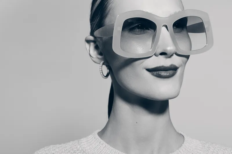 Elegant white woman with pale skin wearing oversized sunglasses and hoop earrings smiles softly in a minimalist studio setting.