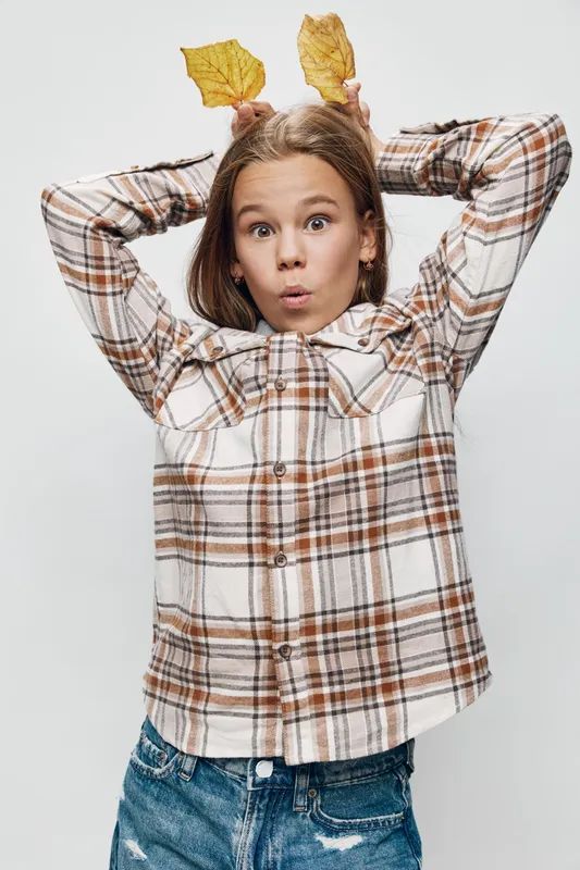 Girl with surprised expression wearing plaid shirt holds autumn leaves behind her head on white background for seasonal fun and playful portrait with casual denim jeans outfit.