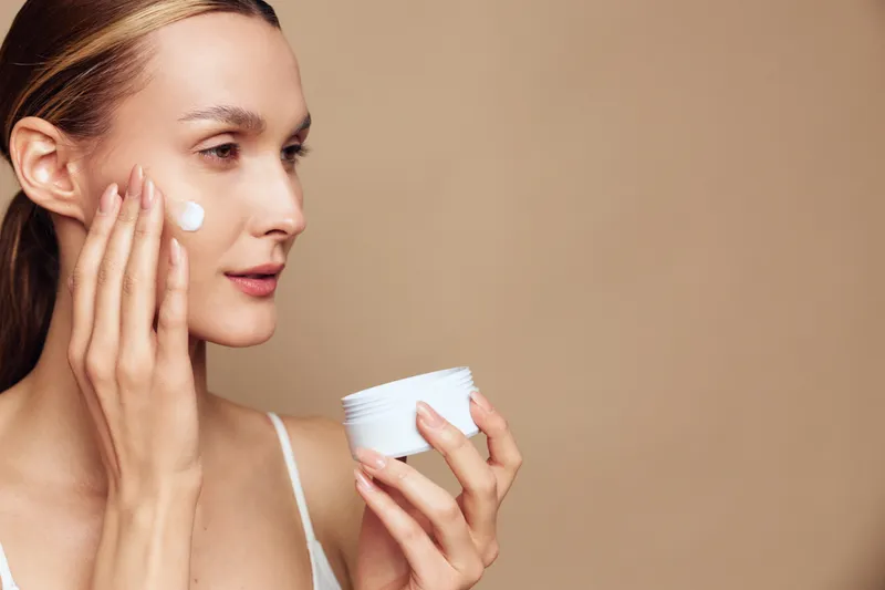 Young woman applies face cream on cheek with fingers holding white jar skincare product isolated on beige background with natural makeup and smooth skin texture.