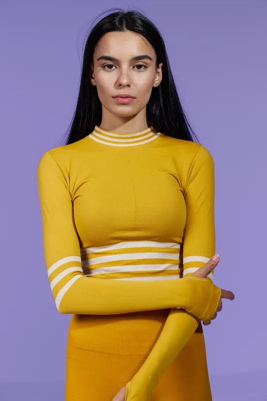 Sport fitness woman wearing yellow athletic top and skirt standing against violet background with focused expression and arms crossed, showcasing strength and determination.