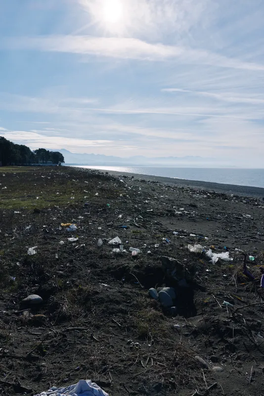 Polluted beach with litter scattered along the shoreline, dark sand and calm water under a cloudy sky, illustrating environmental impact and the need for cleanup and awareness.