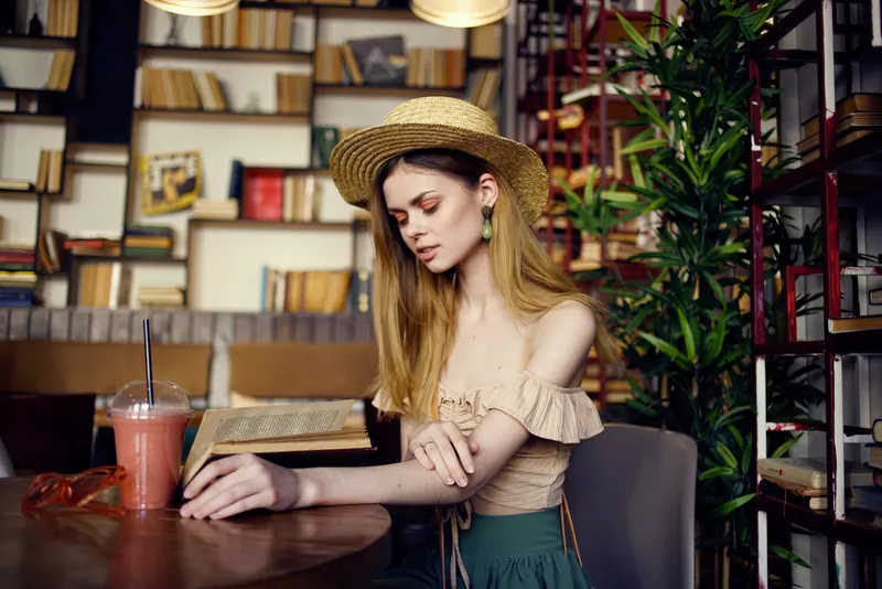 Женщина в шляпе сидит в кафе, у нее на столе напиток и книга, она читает и отдыхает в отпуске