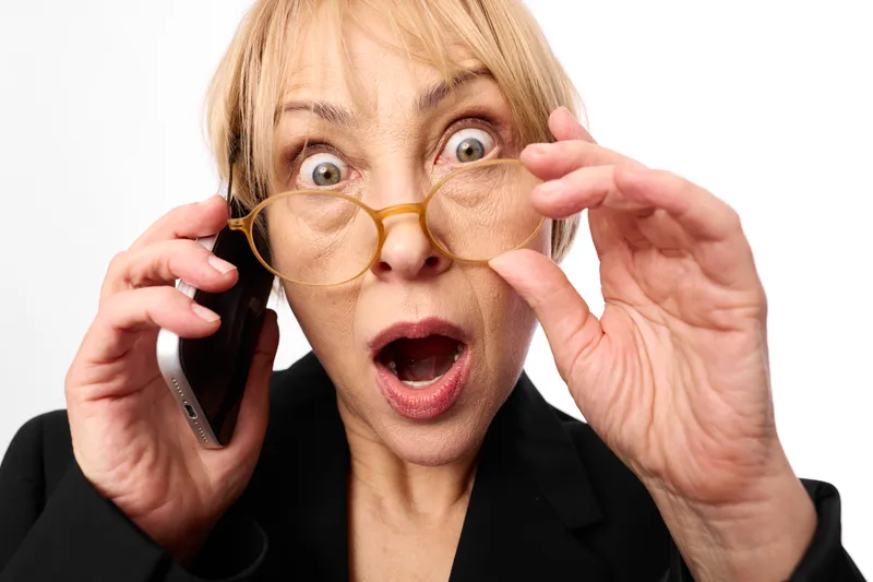 Surprised blonde woman with novelty round glasses, holding smartphone to ear, adjusting glasses with both hands, playful style, studio white background