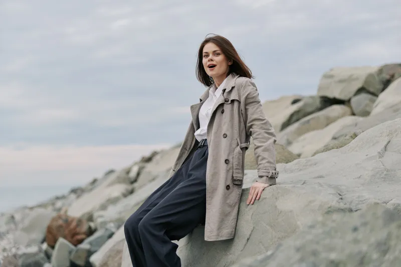 Young woman in beige trench coat sits on large rocks near the coast under cloudy sky. Casual outfit and natural makeup create relaxed outdoor atmosphere by the sea.