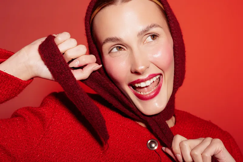 Happy woman in red sweater and headscarf smiles brightly against red background. Close-up portrait with natural makeup and joyful expression. Concept of fashion, beauty, and winter style.