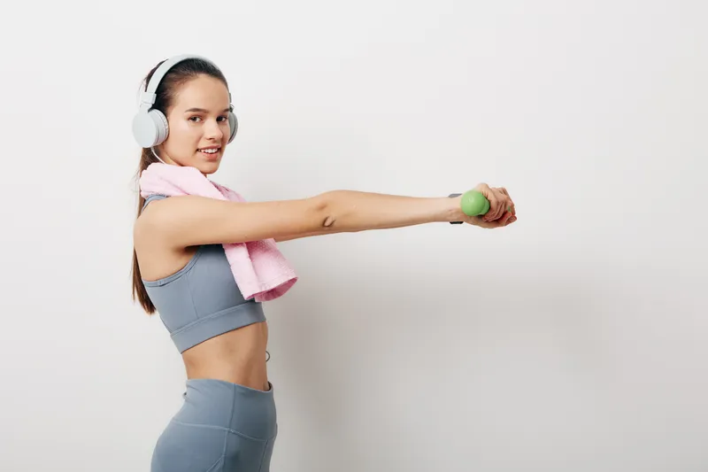 Woman fitness, sport concept: young athletic woman in gray activewear performs a dumbbell lateral raise with white headphones and pink towel on shoulder against a clean white background.