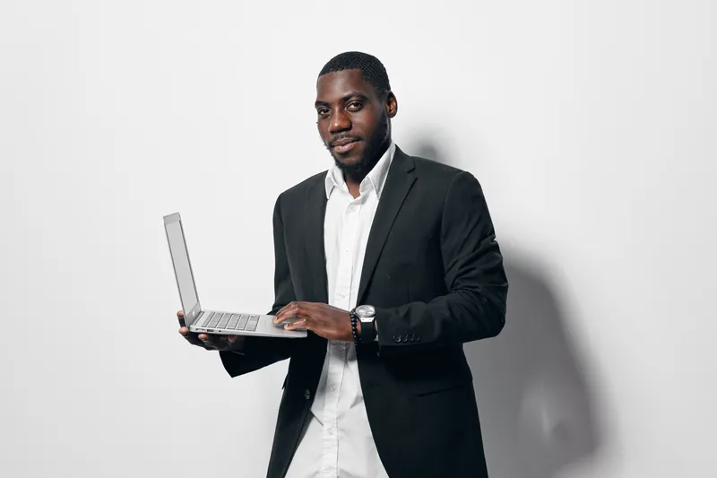 African American man with dark skin wearing black suit and white shirt holding laptop and looking at camera with confident expression on white background in studio.