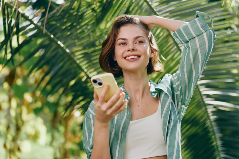 A smiling woman holds a smartphone outdoors among tropical greenery, wearing a striped casual shirt, sunlit and carefree.
