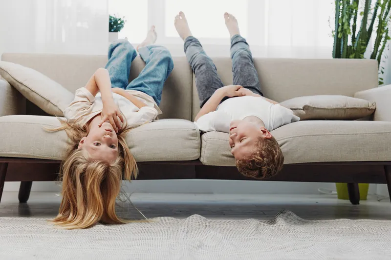 Two children lying upside down on a beige sofa in a bright living room, smiling and relaxed. Casual jeans and white shirts create a playful, cozy home atmosphere with natural light.