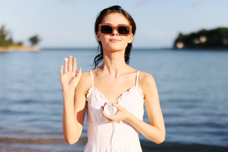 Young woman stands by a calm sea, wearing a pale dress and sunglasses, raising a hand in a friendly wave while a small object rests in her other hand.