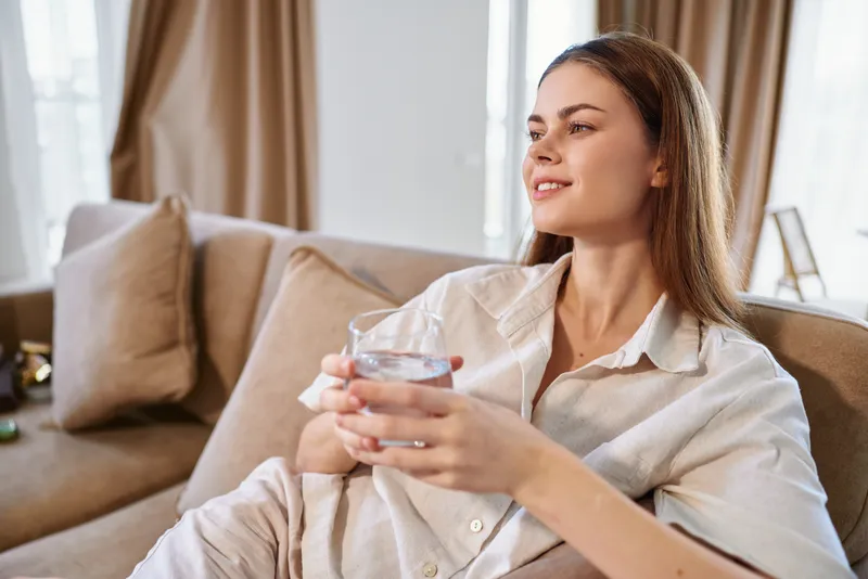 Woman on sofa holding glass of water, relaxed and smiling in cozy home interior. Young female in casual clothes enjoying a quiet moment of comfort and wellness on couch.
