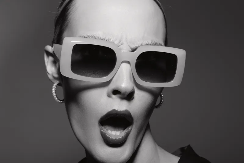 Blonde Caucasian woman with pale skin wears large square sunglasses and pearl earrings, expressing surprise. Studio portrait.