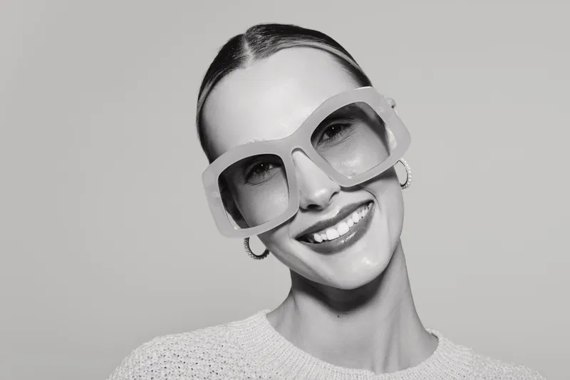 Young white woman with pale skin smiling wearing large sunglasses and hoop earrings on grey background.