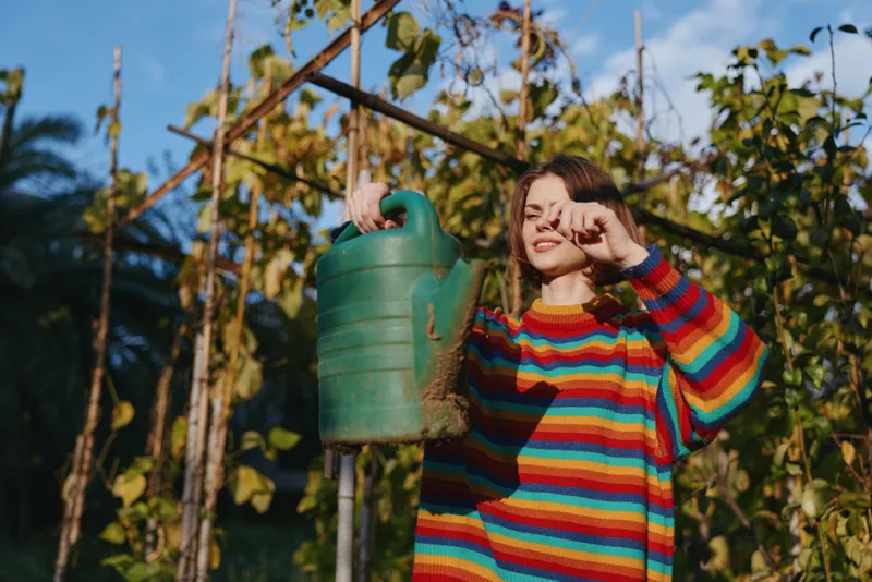 Woman in rainbow sweater watering plants with a green watering can in a tropical garden, young adult smiling in casual outfit, sunny outdoor lifestyle and sustainable gardening hobby.
