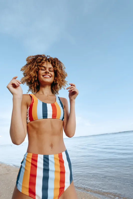 Woman, beach, ocean, swimsuit, curly hair, smiling, sunny sky, outdoors, stands on sandy shore, holds hair near shoulders, relaxed pose, warm daylight, blue water horizon, summer
