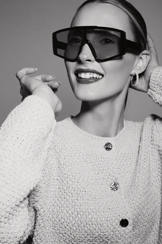 Elegant white woman with fair skin and slicked back hair wears oversized fashion sunglasses and knitted sweater. Studio portrait with confident smile and modern accessories concept.