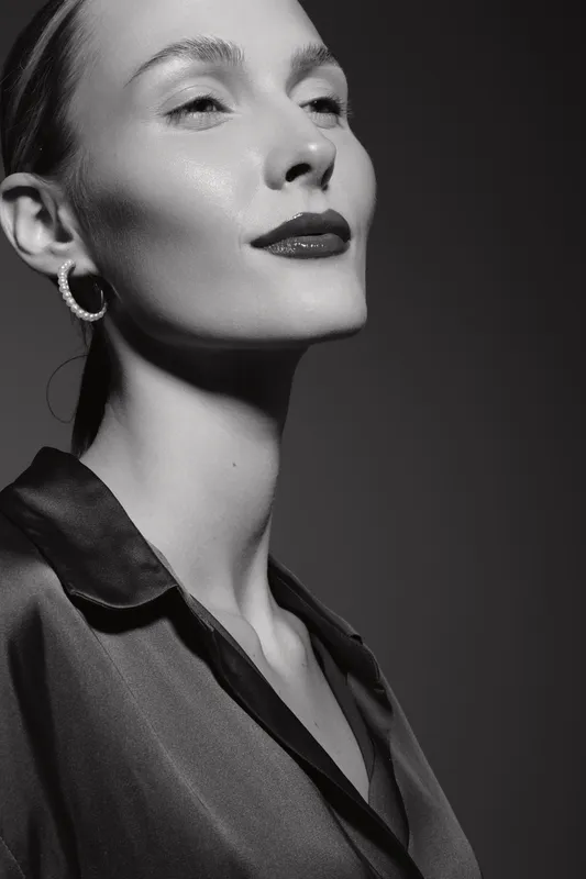 Elegant caucasian woman with pale skin poses confidently in black silk blouse and hoop earrings, studio portrait with dramatic lighting.