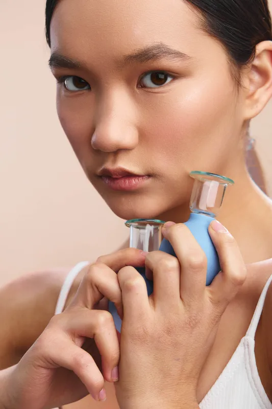 asian woman with a calm expression holds facial massage rollers near her face in a soft studio, highlighting a skincare routine and a serene self care moment.