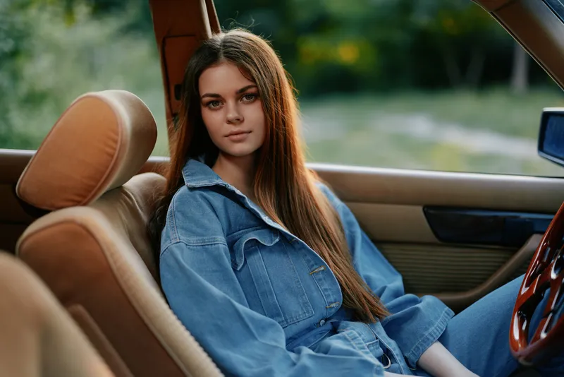 Young woman in denim outfit sitting in a vintage car, displaying a relaxed emotion in soft natural light surrounded by greenery. Perfect for lifestyle and fashion themes.
