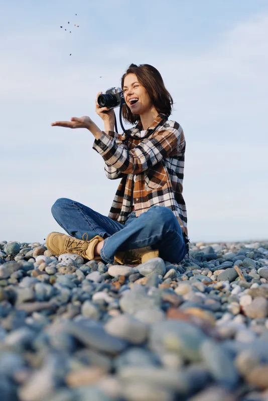 Woman photographer on a rocky beach, smiling while shooting with a compact camera, wearing a plaid shirt and jeans, enjoying a sunny day and capturing candid moments by the shore