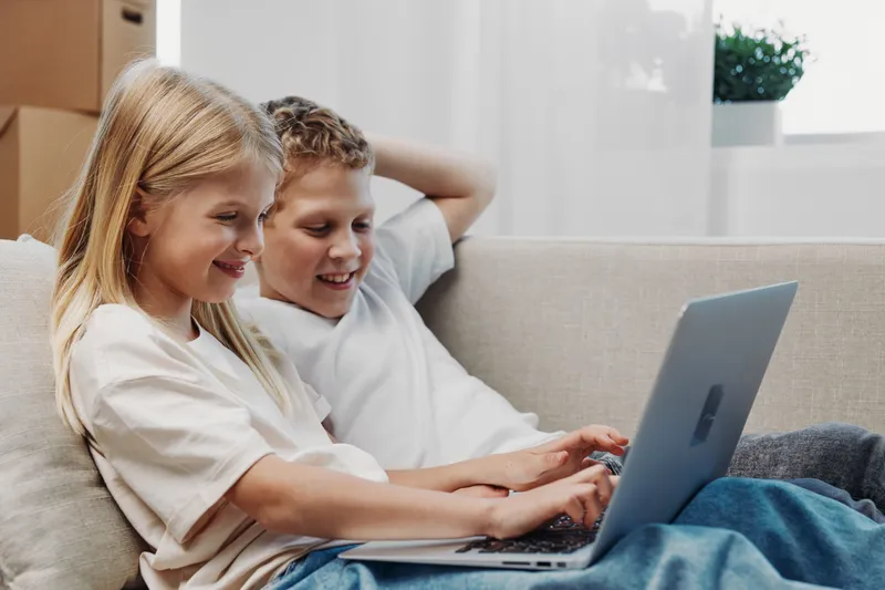 Smiling children using laptop on couch in bright living room, enjoying technology and learning together at home. Concept of education and digital connection.