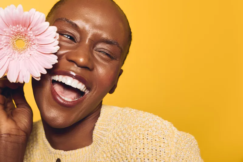Happy African woman with dark skin smiles widely holding pink flower near face on yellow background. Concept of joy, beauty, natural skincare, and positive emotions in studio lighting.