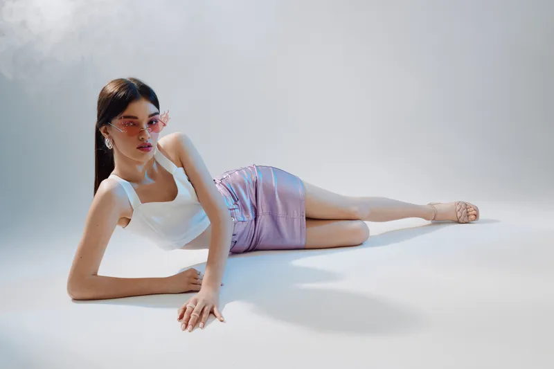 Young woman in white top and pink skirt poses lying on floor with stylish sunglasses. Fashion concept focused on beauty, elegance, and modern style in studio lighting.