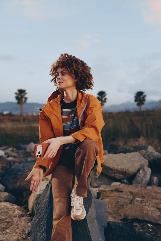 Woman lifestyle outdoors, curly hair, orange jacket, seated on rocks, striped shirt, soft natural light, cloudy sky, looking at camera, calm travel mood in scenic landscape