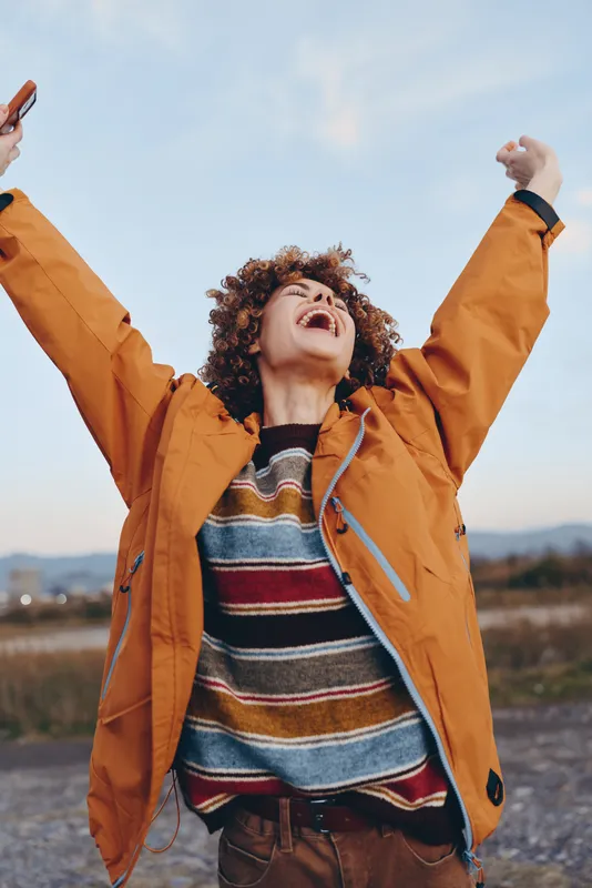 Woman with curly hair in orange jacket outdoors, laughing with arms raised under open sky, lifestyle moment of joyful freedom, casual fashion and bright light